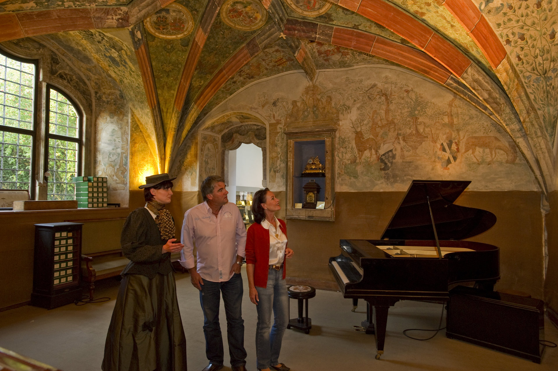 Ein Paar wird von einer Dame im Gewand durch das Museum geführt. In einem schön ausgeleuchteten Raum steht ein Klavier.