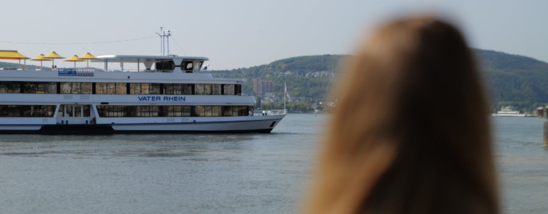 Im Vordergrund ist unscharf der Rücken einer Frau zu erkennen, die auf ein großes Ausflugsschiff auf dem Rhein blickt. Das weiße Schiff mit dem Namen „Vater Rhein“ fährt auf dem Fluss, im Hintergrund sind grüne Hügel und ein paar Gebäude zu erkennen. 