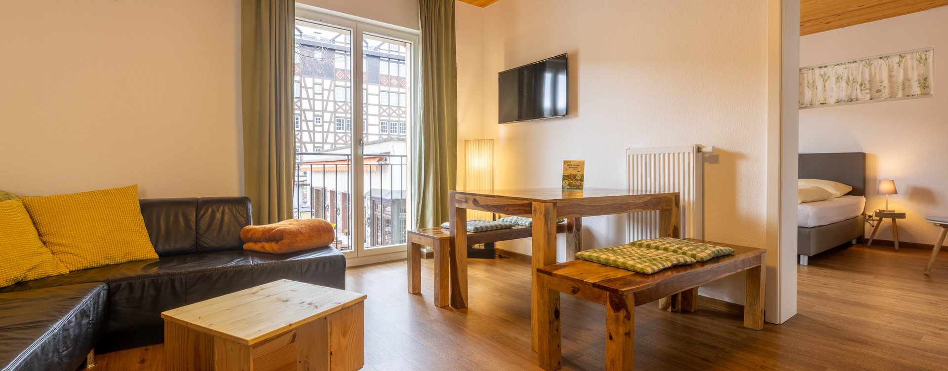 Helle Ferienwohnung Querfeldheim in Rüdesheim mit gemütlichem Wohnbereich, Holzmöbeln, schwarzer Ledercouch und Blick auf Fachwerkhäuser.