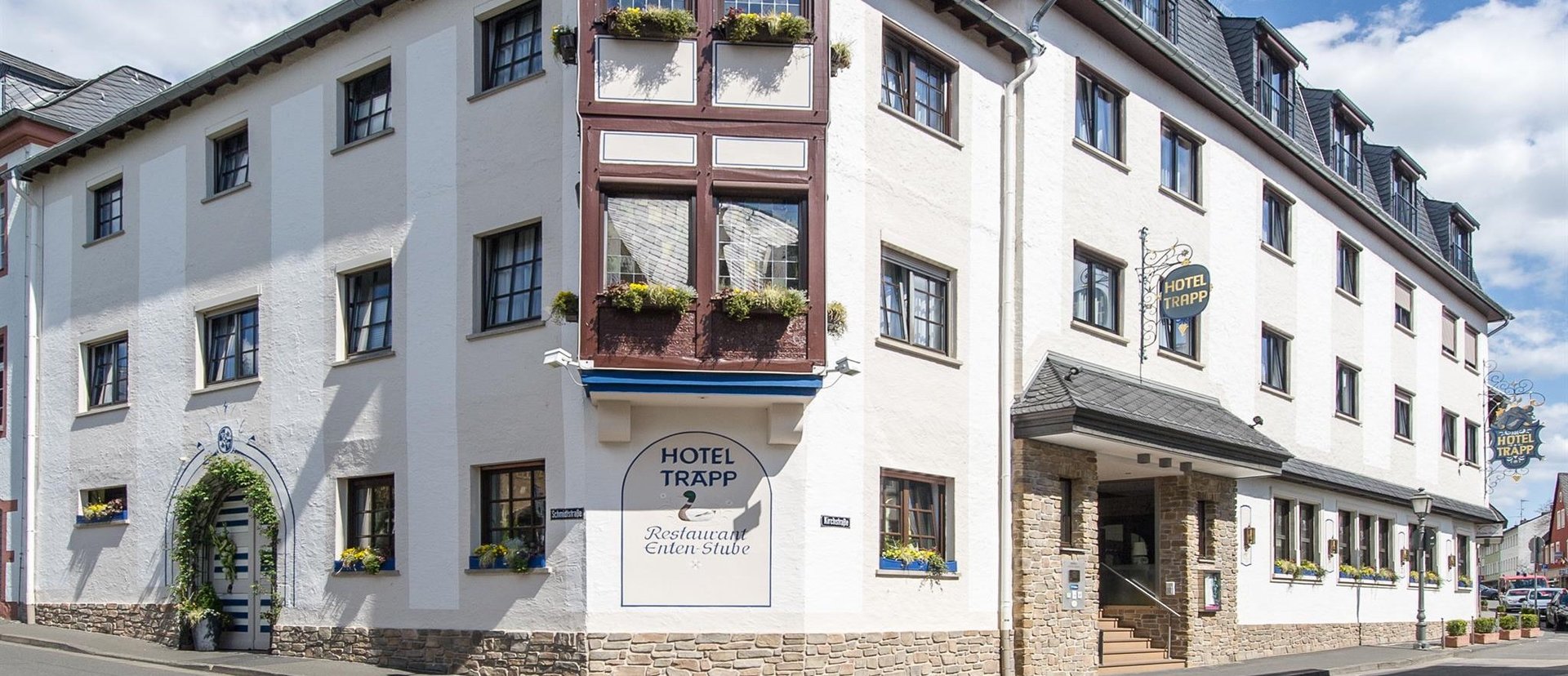 Ansicht des Hotels Trapp, ein großes, weißes Gebäude mit mehreren Fenstern und einem dekorativen Fensterbereich mit Blumenkästen. Das Hotel liegt an einer Straßenecke unter blauem Himmel.
