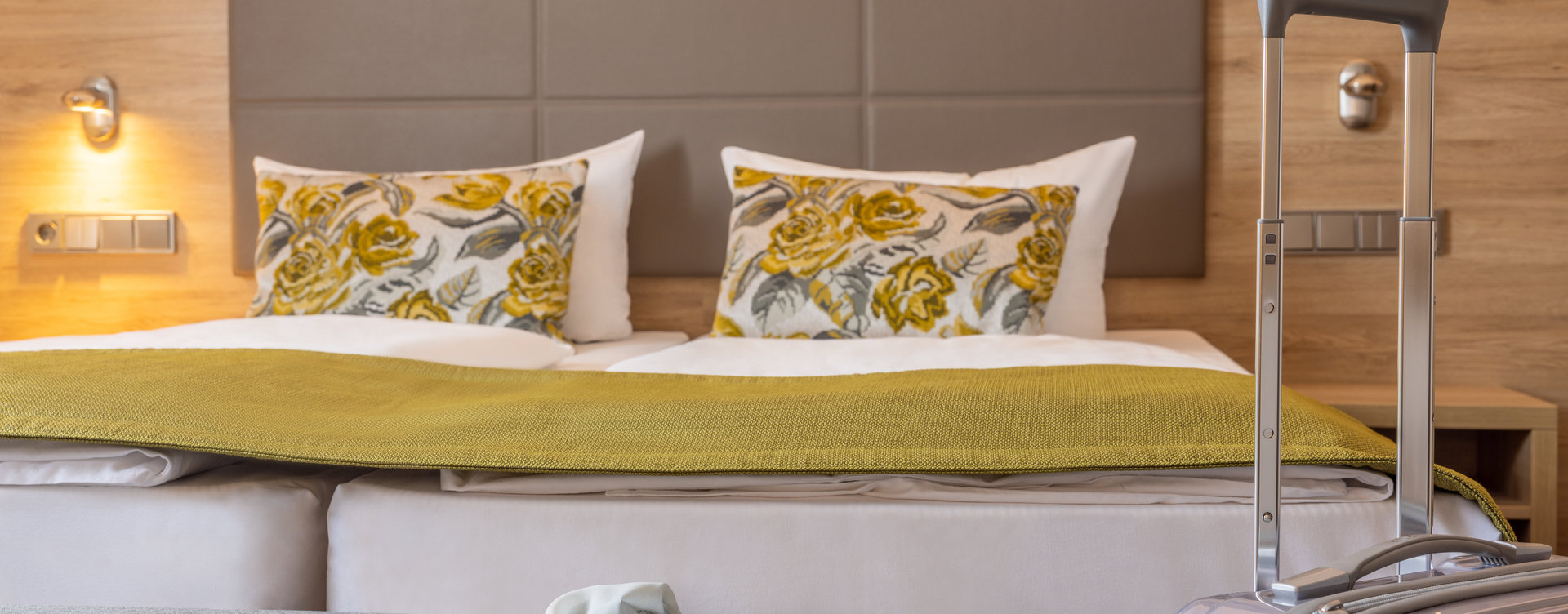 Hotelzimmer mit einem großen Doppelbett, dekorativen Kissen und einer Tagesdecke. Vor dem Bett stehen eine graue Bank und ein silberner Koffer.