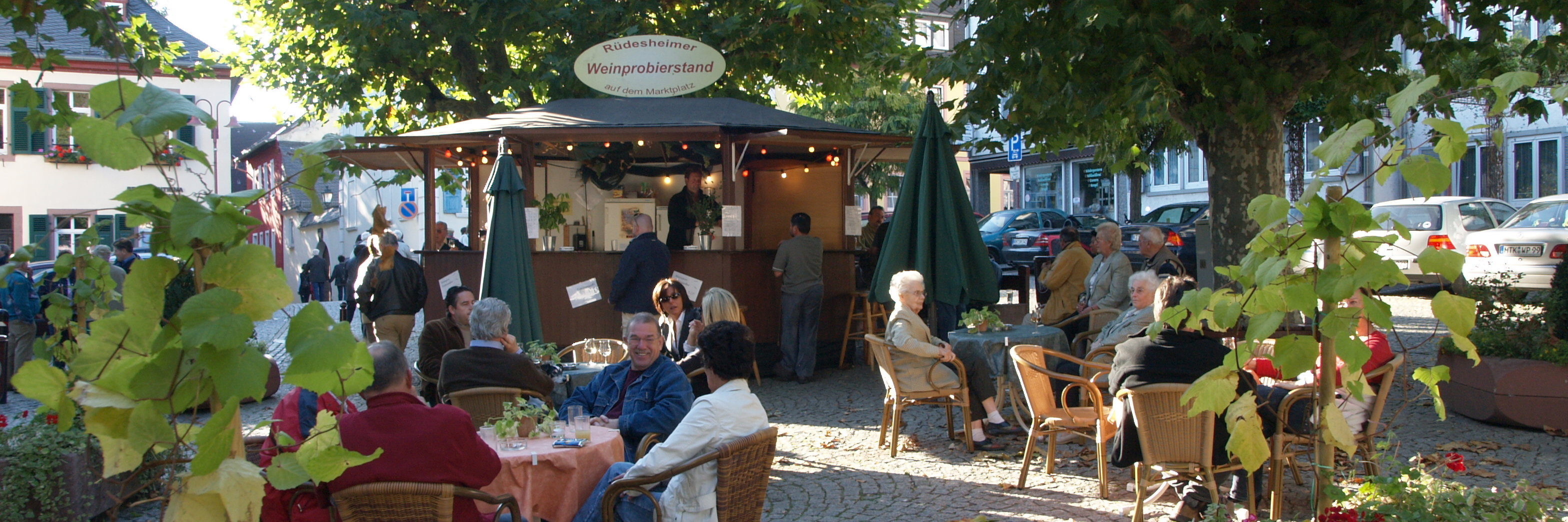 Von Bäumen umgeben staht ein Weinstand auf dem Marktplatz in Rüdesheim. In gemütlicher Atmosphäre trinken viele Personen an Tischen sitzend und am Weinstand Wein.