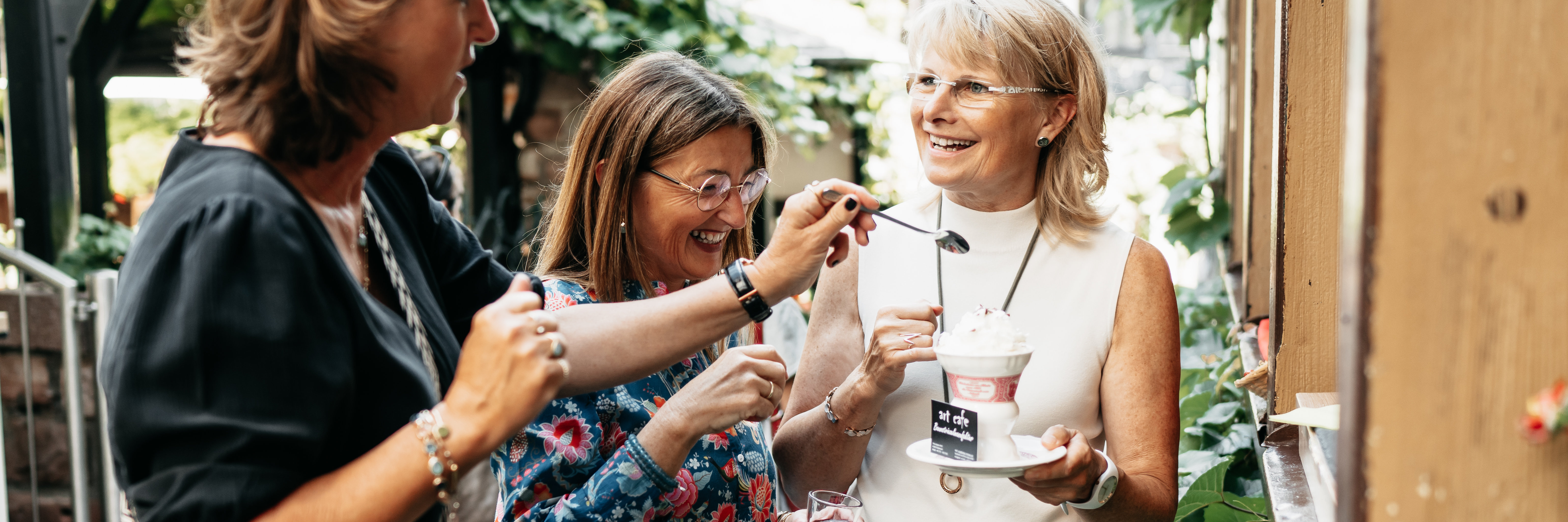 Drei Frauen stehen draußen in der Drosselgasse. Die Frau ganz rechts hält einen Rüdesheimer Kaffee in der Hand. Die Frauen lachen sich gegenseitig an. 