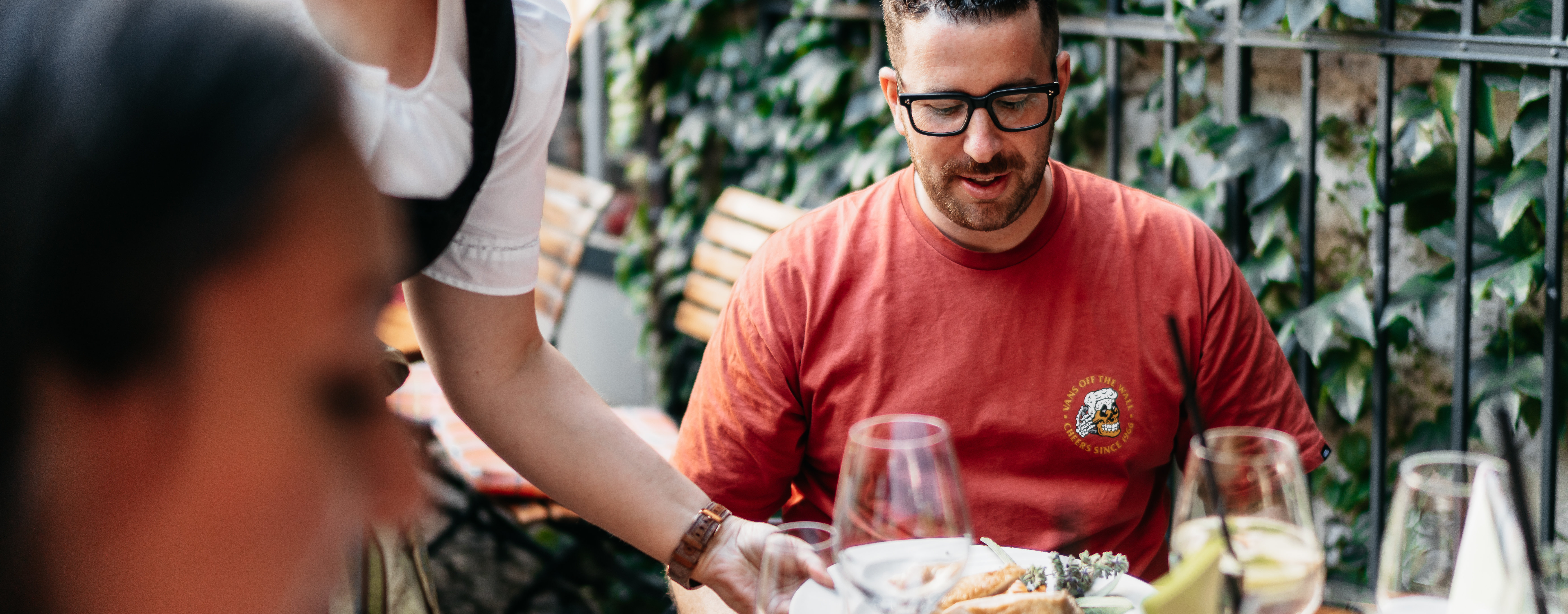 Ein Mann sitzt in einem Restaurant in der Drosselgasse. Eine Frau die als Servicekraft arbeitet bedient den Mann und bringt ihm sein bestelltes Essen an den Tisch.