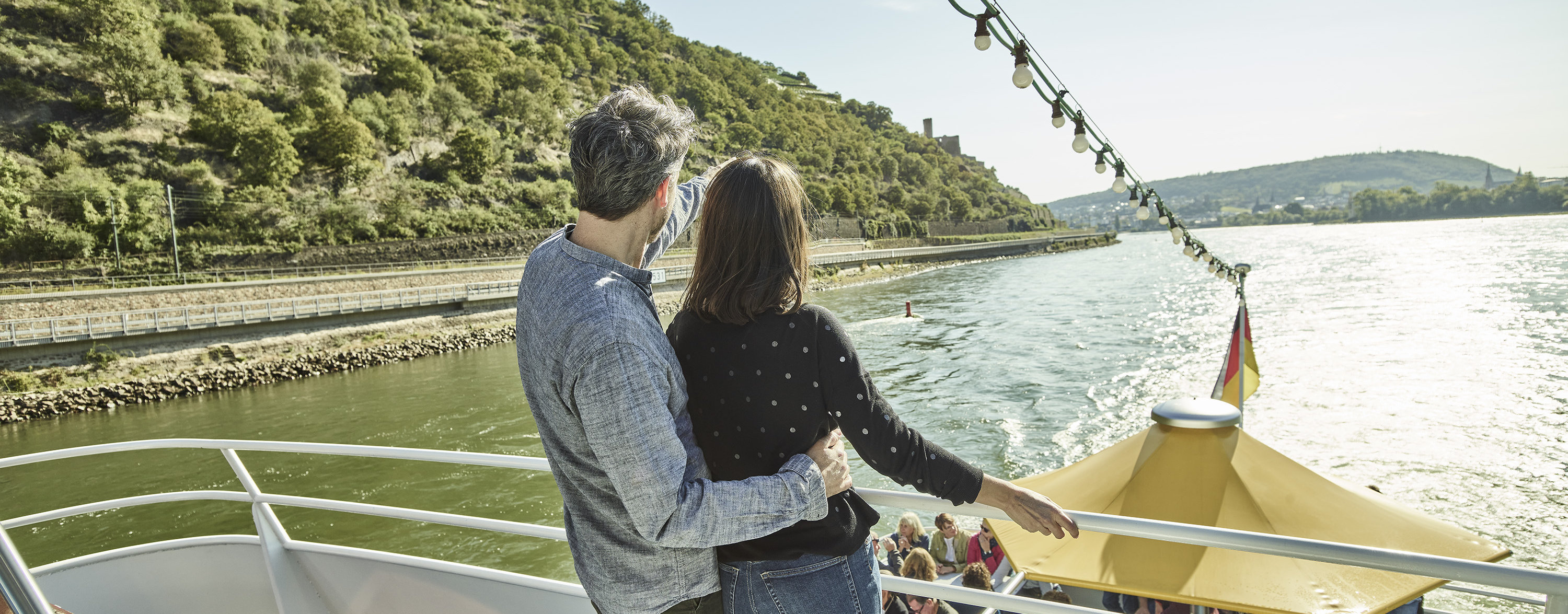 Ein Paar steht bei Sonnenschein auf einem Schiff und blickt auf das Wasser und die Weinberge.
