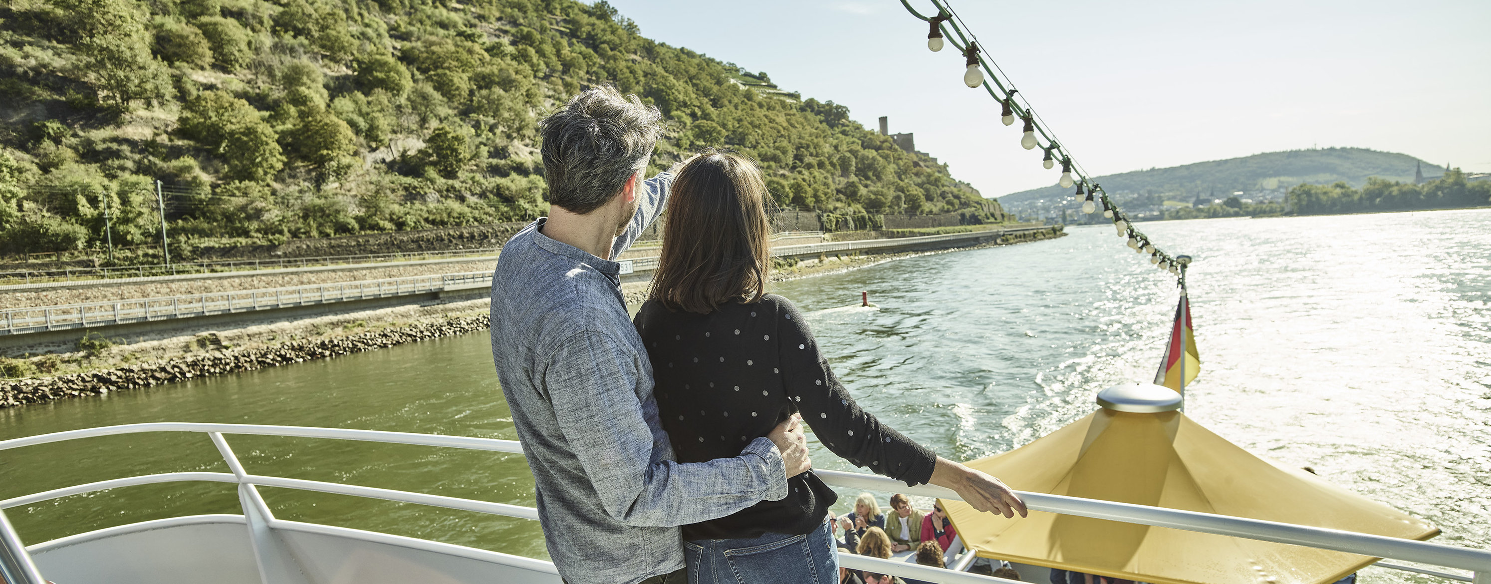 Ein Paar steht bei Sonnenschein auf einem Schiff und blickt auf das Wasser und die Weinberge.