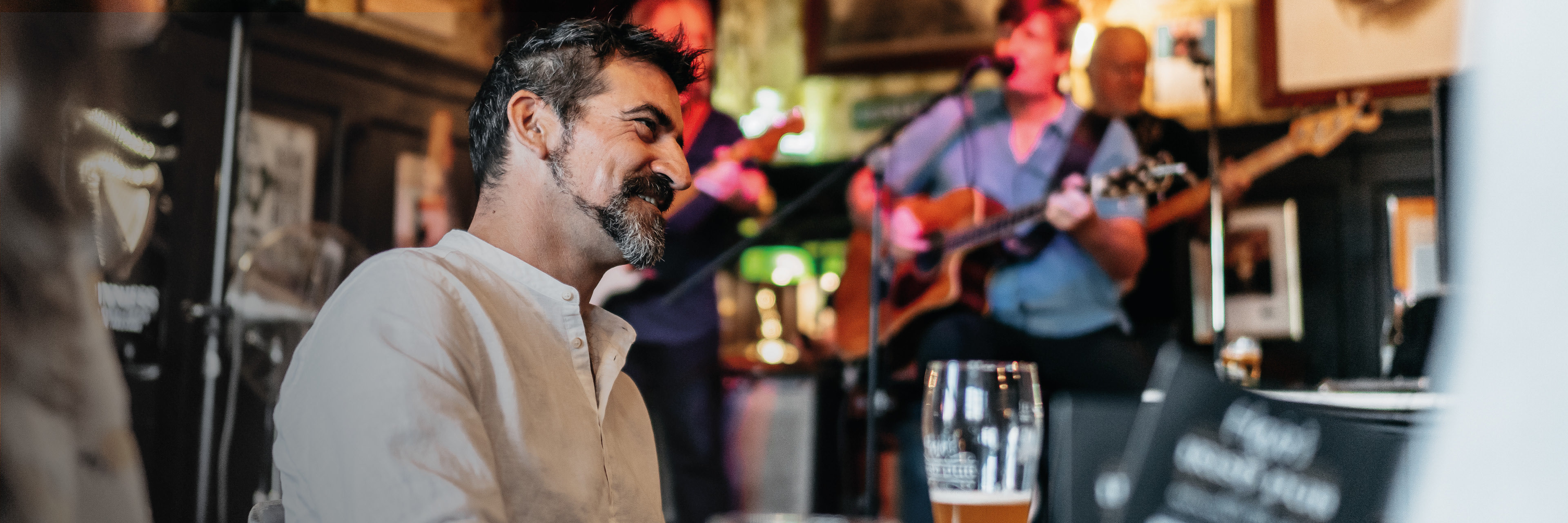 Ein Mann sitzt in einer Kneipe mit einem Glas Bier in der Hand am Tisch und im Hintergrund spielt eine Band.