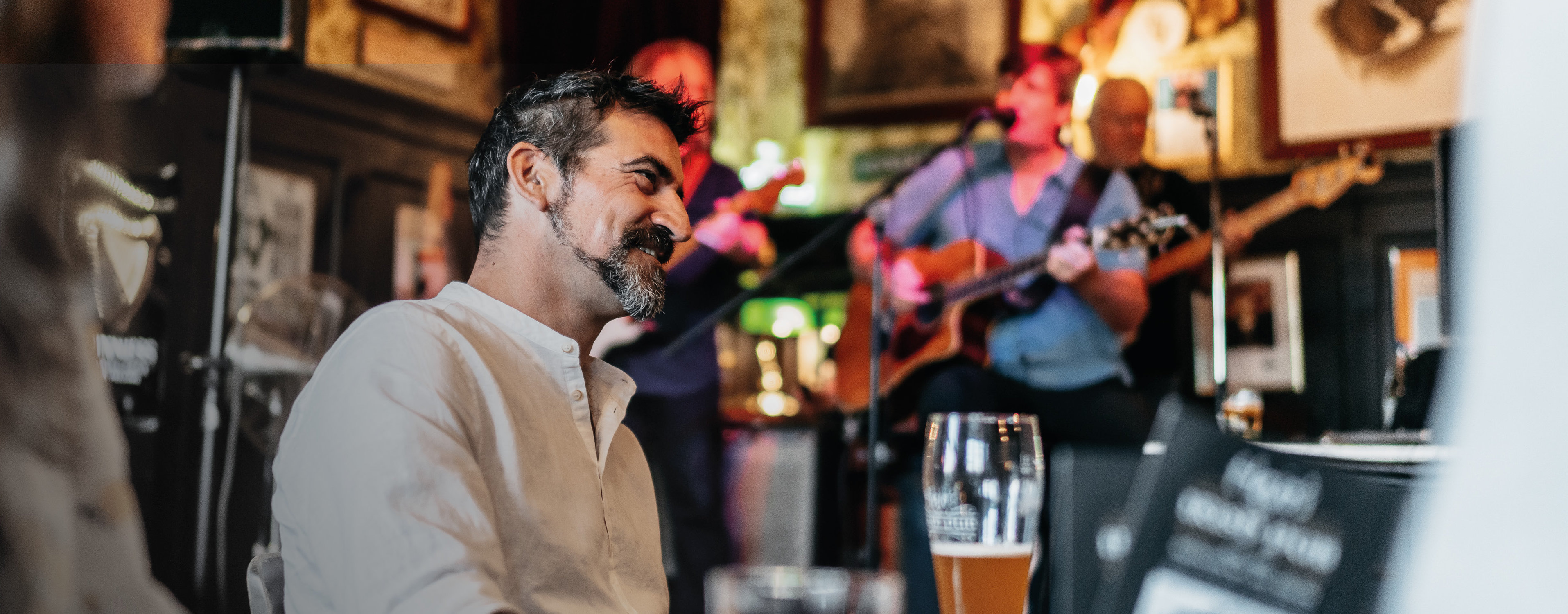 Ein Mann sitzt in einer Kneipe mit einem Glas Bier in der Hand am Tisch und im Hintergrund spielt eine Band.