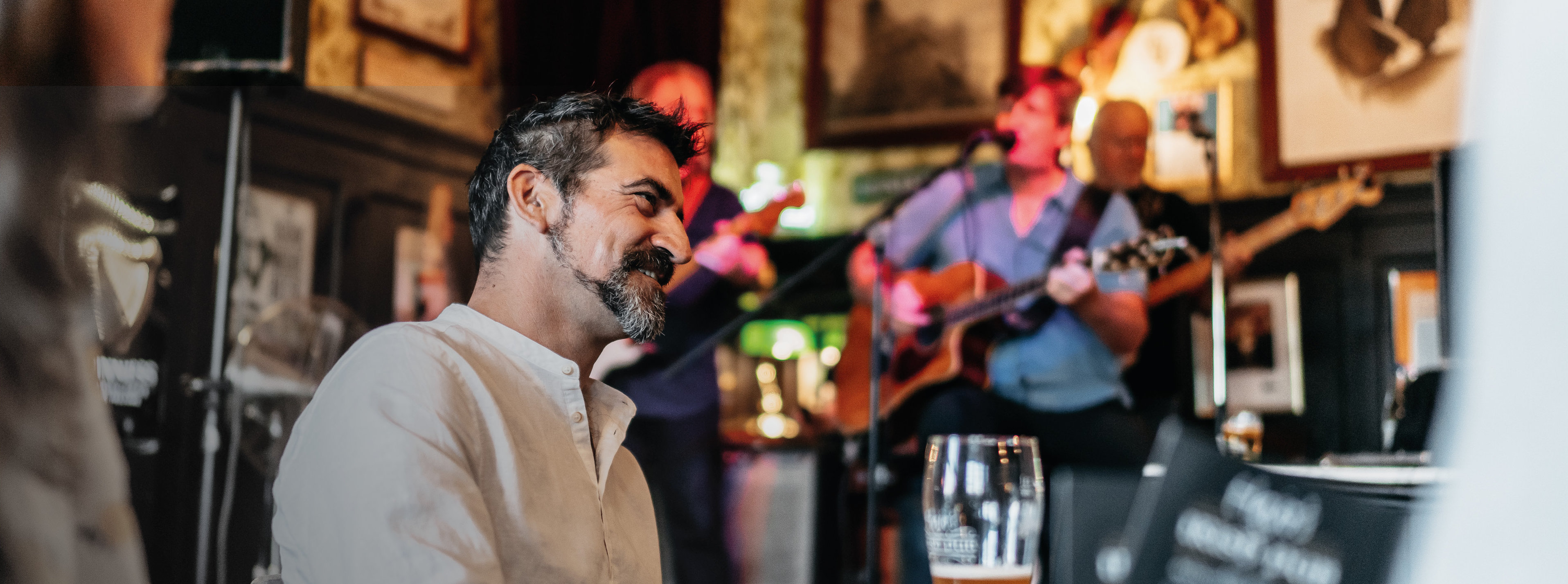 Ein Mann sitzt in einer Kneipe mit einem Glas Bier in der Hand am Tisch und im Hintergrund spielt eine Band.