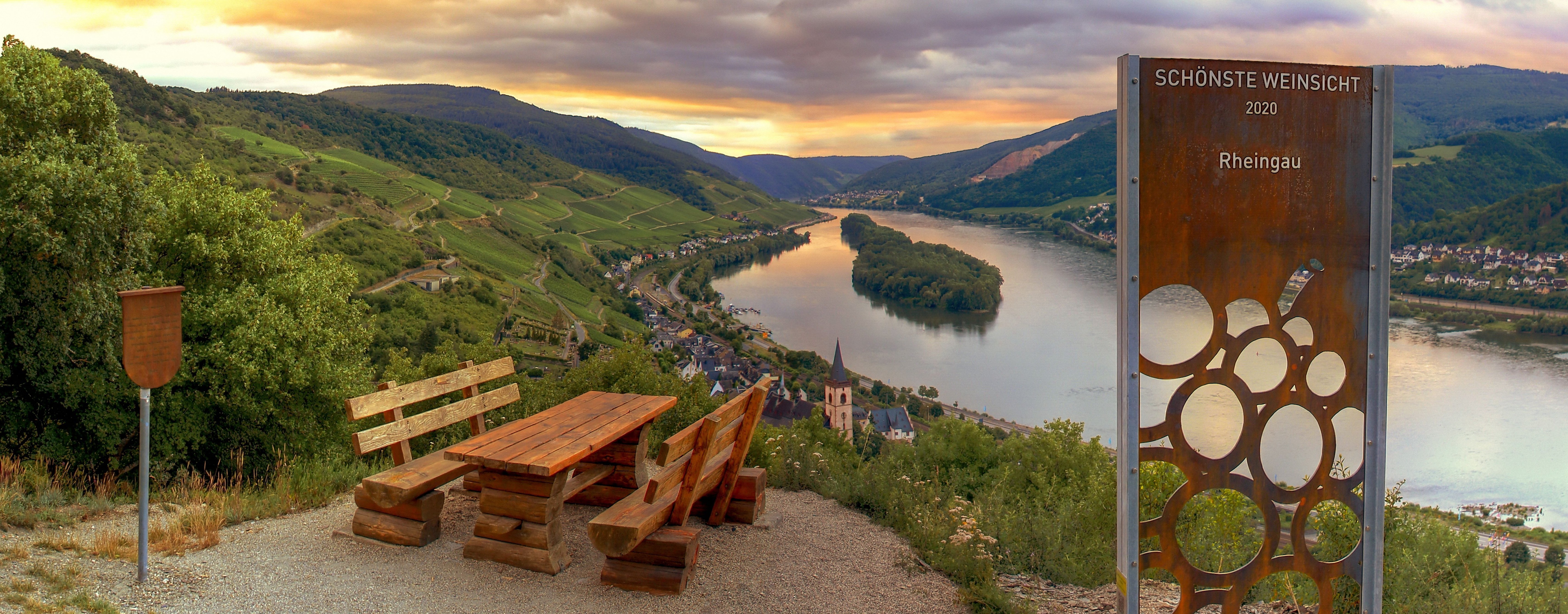 Bei Sonnenuntergang blickt man von einem Hügel auf die Weinberge und den Rhein. Am Ausblickspunkt ist ein Rastplatz mit Tisch und Bänken.