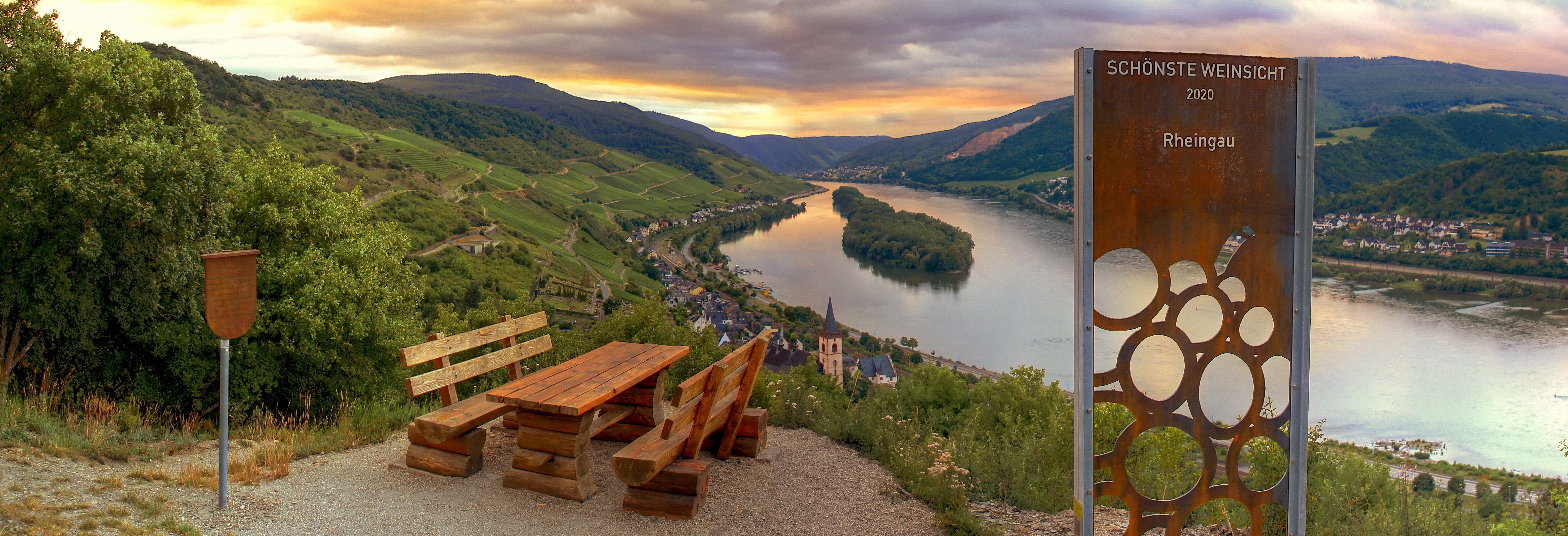 Bei Sonnenuntergang blickt man von einem Hügel auf die Weinberge und den Rhein. Am Ausblickspunkt ist ein Rastplatz mit Tisch und Bänken.