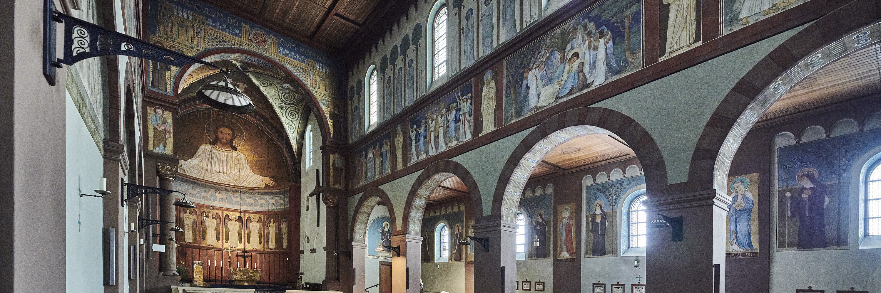 Innenraum der Abteikirche St. Hildegard in Rüdesheim, Blick auf den Altarraum mit großer Christus-Darstellung an der Wand. Die Kirche ist mit Holzdecken, Wandmalereien, Rundbögen und Kirchenbänken gestaltet.