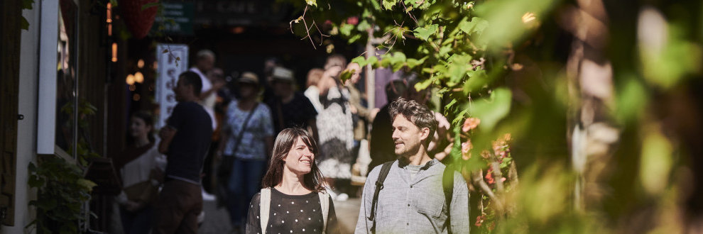 Ein Paar geht Händchenhaltend und lachend durch die engen Gassen der Altstadt von Rüdesheim am Rhein. Im Hintergrund sind viele Menschen verschwommen zu sehen.