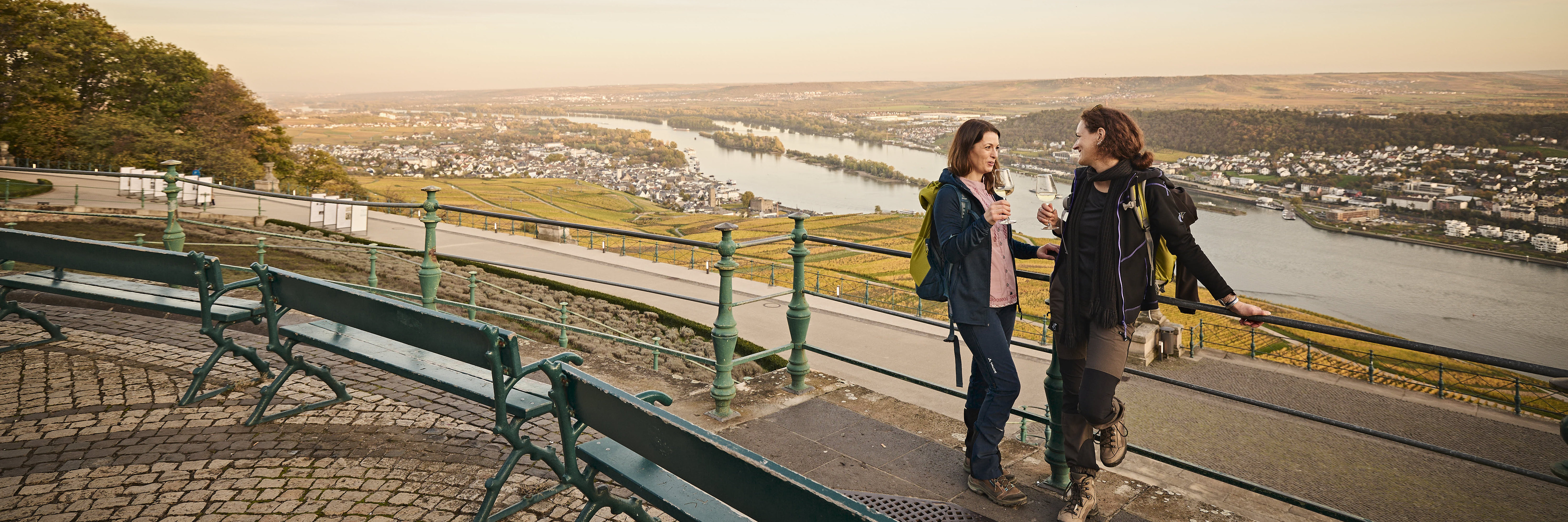 Zwei Damen stehen in Wanderkleidung unterhalb der Germania, blicken diese an und haben jeweils ein Glas Wein in der Hand. 