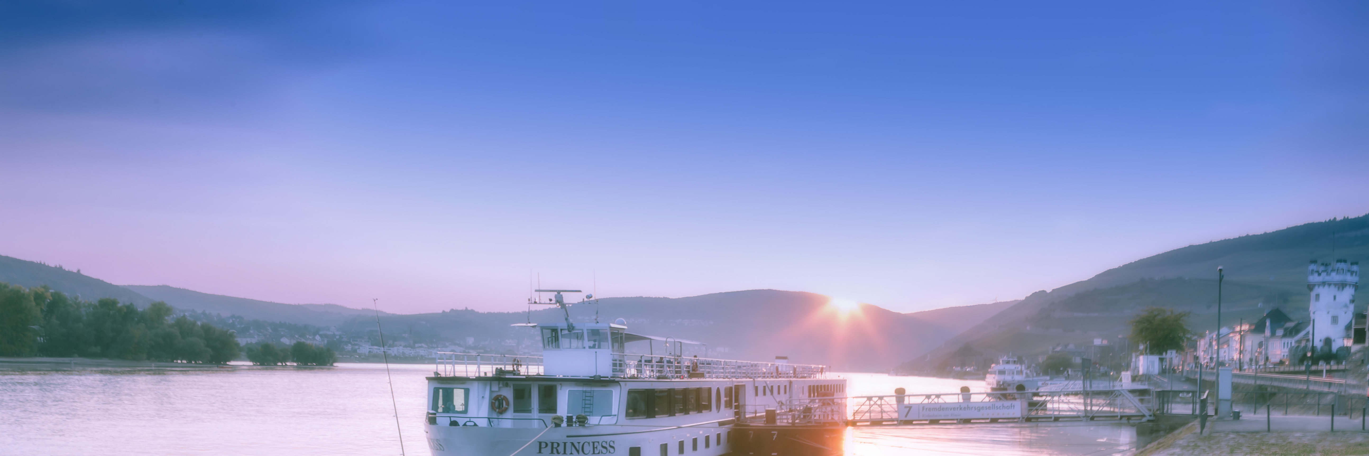 Das Bild zeigt die Uferpromenade von Rüdesheim am Rhein in der Abenddämmerung. Im Vordergrund ist ein Anlegesteg mit einem festgemachten Passagierschiff zu sehen, auf dem der Name „Princess“ steht.  Auf der rechten Seite erkennt man Teile der Altstadt und den Adlerturm.