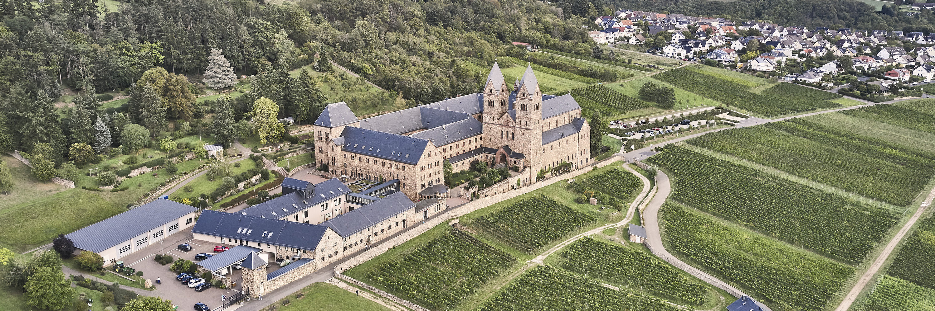 Die Luftaufnahme der Abtei St. Hildegard zeigt das Kloster von oben umgeben von Weinbergen bei leicht bewölktem Himmel