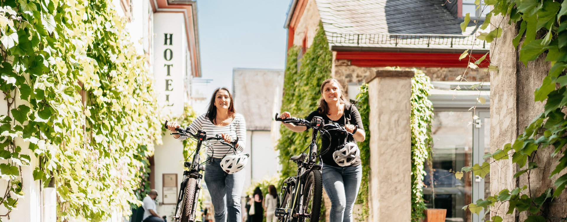 Zwei Frauen schieben Ihre Fahrräder durch die Drosselgasse in Rüdesheim. Es ist sommerliches Wetter. 