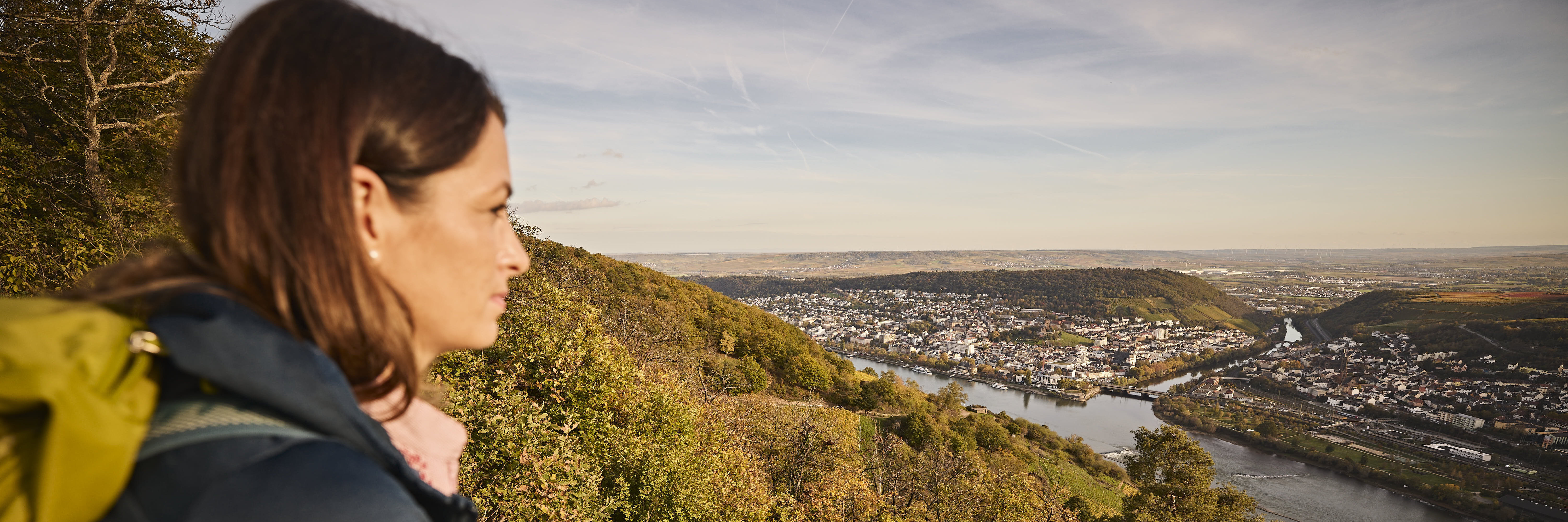 Eine Frau lehnt an einer Steinmauer und blickt auf den Rhein, umgeben von grünen Hügeln, mit der Stadt Bingen am gegenüberliegenden Ufer bei sonnigem Himmel.