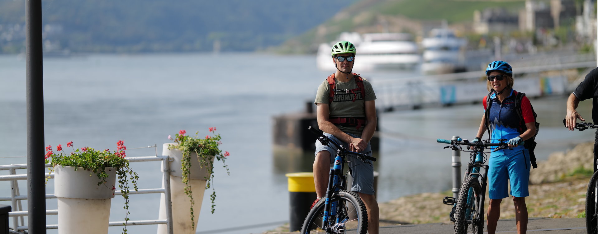 Ein Paar am Rhein macht eine Pause an der Rüdesheimer Rheinpromenade. Der Mann sitzt auf seinem Fahrradsitz und die Frau steht daneben und hält Ihr Fahrrad am Lenkrad fest. Im Hintergrund sieht man den Rhein, die Weinberge und zwei Schiffe.
