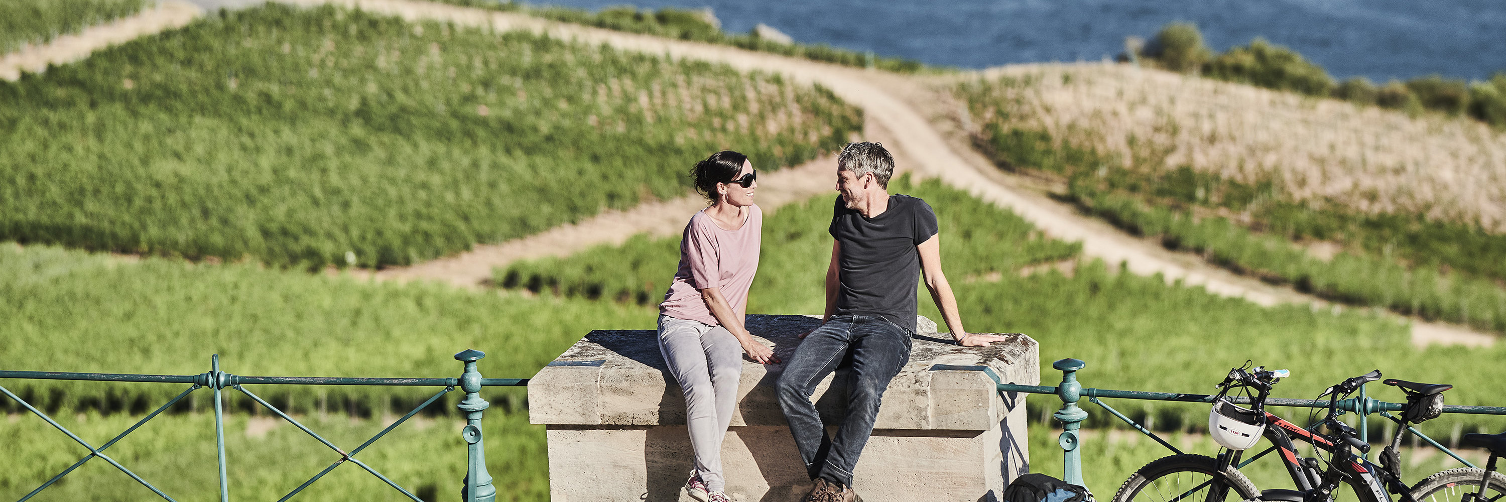 Ein Paar macht während einer Radtour eine Pause beim Niderwalddenkmal. Sie sitzen gemütlich auf einem Stein und haben die Aussicht auf den Rhein und die Weinberge.